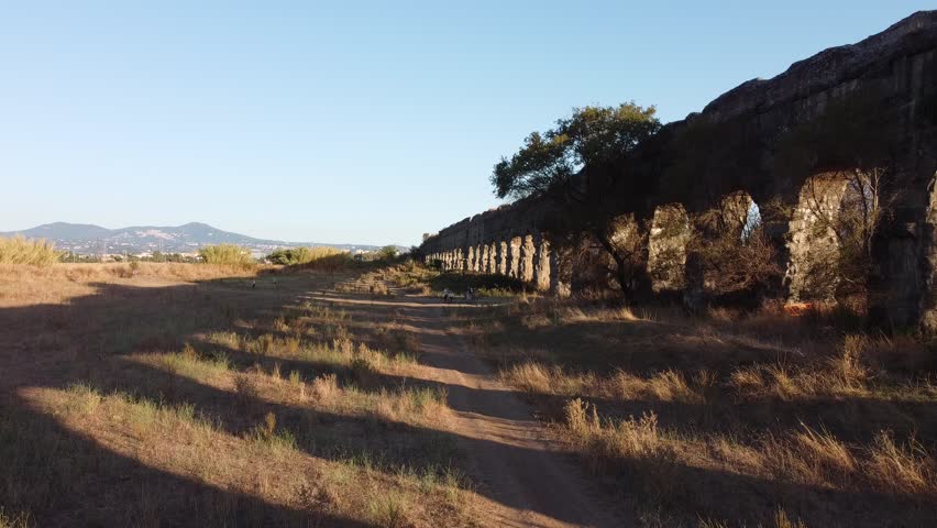 Drone aerial view of an ancient Roman aqueduct crossing a dry rural landscape at sunset. Historic stone arches extend across the countryside with mountains and fields in the background under warm golden light.