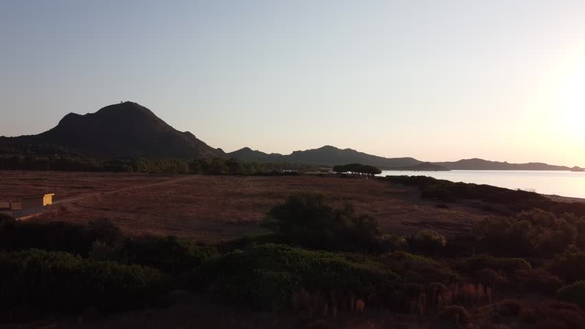 Drone aerial view of a long natural sandy coastline with gentle waves and calm sea during sunset. Scenic coastal landscape with soft evening light and distant mountains along the horizon.