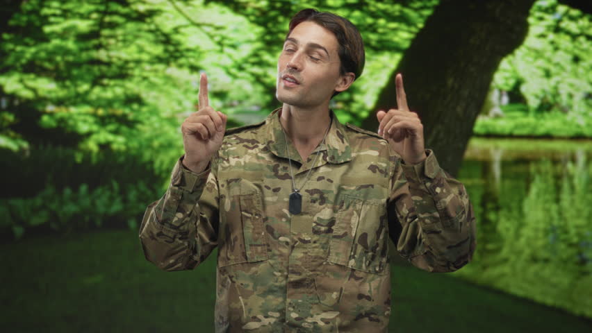 Soldier man in camouflage jacket shows dogtags and gives two thumbs up beside a pond in forest; pride duty service.