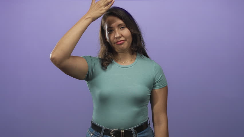 Young woman in teal shirt smiling with hand across mouth gesture in purple studio; playful confidence.