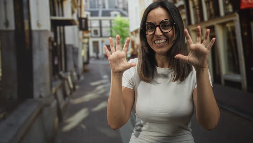 Woman smiling with hands raised, fingers splayed making playful claw gesture in narrow street; joy playfulness.