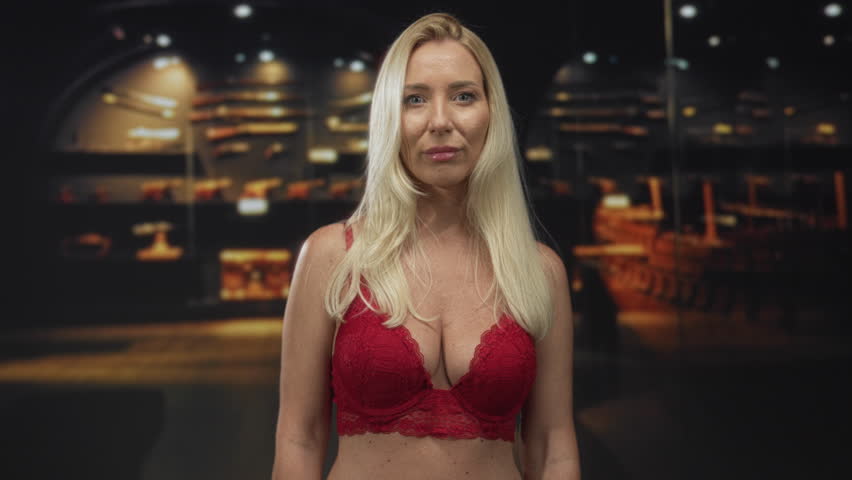 Woman with blonde hair wearing red lace bra exposing chest standing in museum building gazing ahead; confidence.