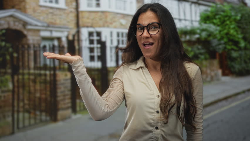 Young hispanic woman smiling with palm up presenting at house entrance on street, wearing glasses and beige shirt; welcoming warmth.