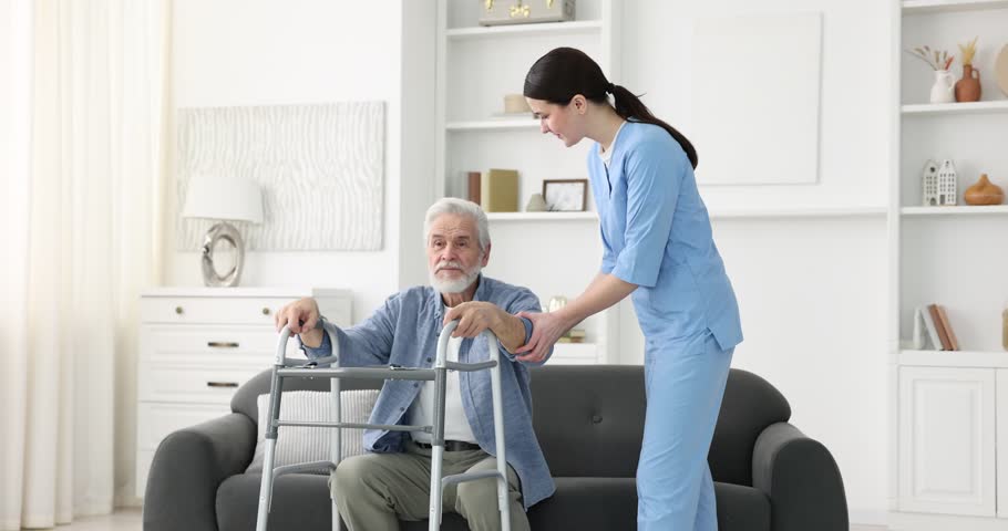 Care worker helping senior man with walker to stand up from sofa indoors