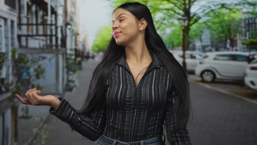 Young latina woman pointing thumbs to chest while tilting her head and smiling on a sunlit city street; confidence.