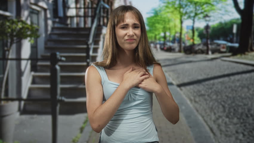 Young woman clutching chest for heartattack on a city street wearing light blue tank top and holding both hands to sternum; distress.