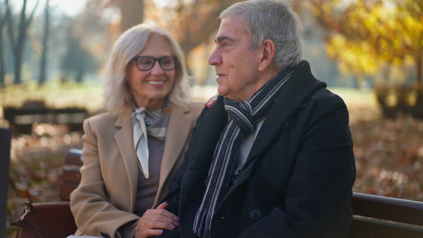 A smiling senior man and woman are sitting close on a wooden bench in a park, with colorful fall foliage in the background