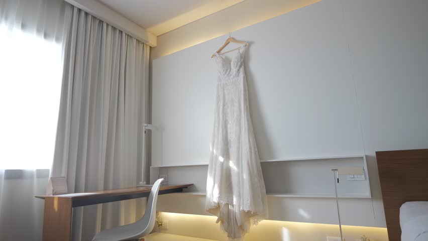 A white dress is hanging on a clothesline in a hotel room. The dress is long and flowing, and it is hanging from a clothesline on the wall. The room is clean and well-lit, with a desk