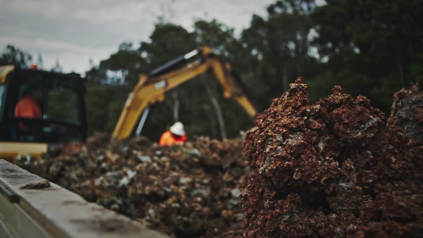 Close-up of freshly excavated red soil in sharp focus as an excavator operates in the background within a graveyard.