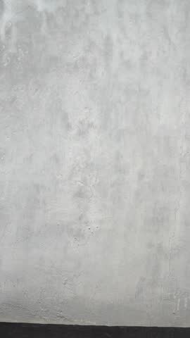 Asian man in a brown shirt walking slowly across the frame while looking down. Authentic real people footage featuring a textured grey concrete wall background with massive copy space. Shot in 4K resolution.