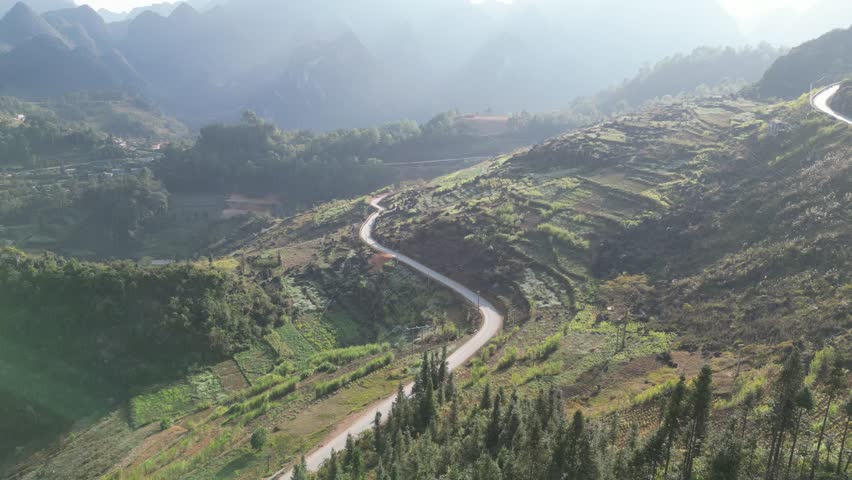 Drone video reveals a stunning aerial view of the winding mountain roads and terraced fields near Mã Pí Lèng Pass, Hà Giang, Vietnam. The landscape captures the sunlit, lush green hills in breathtakin