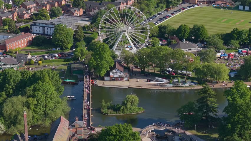 A picturesque static aerial shot of Stratford-upon-Avon in spring. Features the Clopton Bridge, a passing boat on the River Avon, and the giant Ferris wheel surrounded by blooming blossoms.