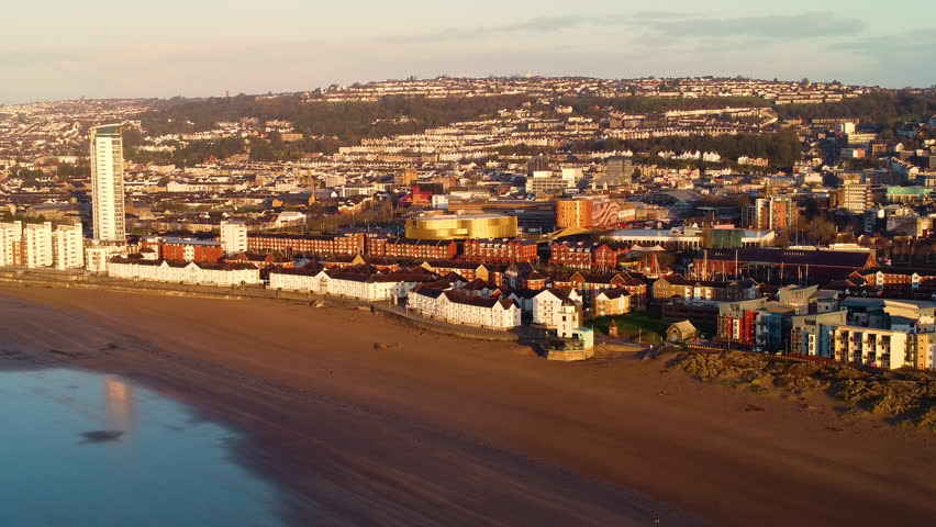 Pushing Aerial of Swansea City Centre with Tall Buildings and Arena with Houses on Hillside. Sunrise Drone of Cityscape in South Wales, UK. Seaside Urban Cityscape Environment.