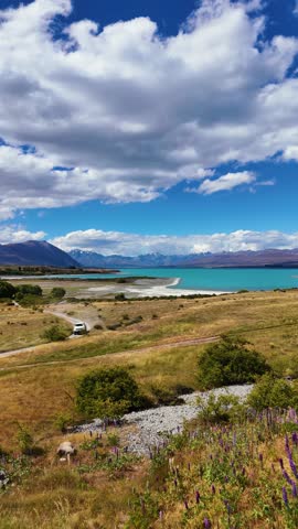 Summer panorama of Lake Tekapo’s turquoise shoreline with winding road, golden tussock hills and the Southern Alps under dramatic clouds, filmed vertical.