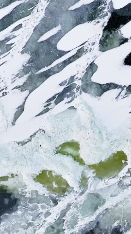 Vertical aerial close-up of frozen sea textures with crushed ice, snow crust, translucent slabs, and green algae patches forming abstract winter patterns in polar water.