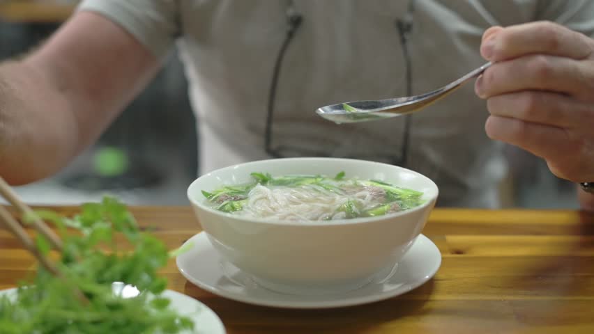 Bowl Vietnamese Pho Bo Steaming On Wooden Table, Caucasian Traveler Samples Clear Beef Broth With Spoon And Chopsticks, Fresh Cilantro And Herbs On Side, Casual Street Cafe Atmosphere