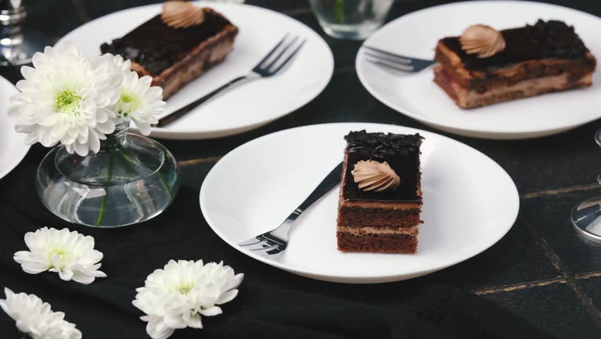 Three chocolate cakes with cream on white plates with forks near white flowers and black fabric