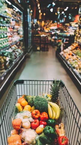 Shopping cart full of fresh fruit and vegetable, products inside a supermarket aisle, healthy lifestyle and grocery shopping concept. Eco market, shop. Vertical pov view.