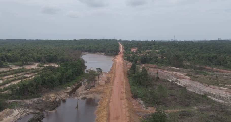 Forward aerial red car driving on a long dirt road between a lake and dense forest, 
