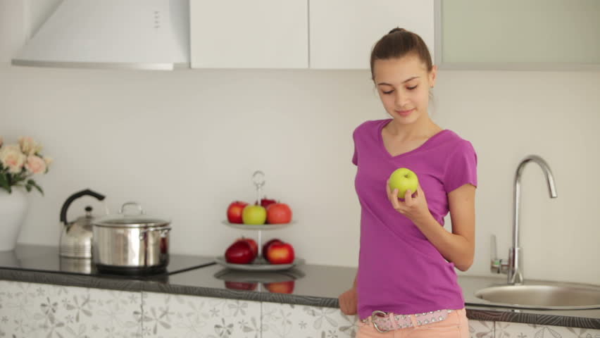 Cute girl is standing in the kitchen and eating an apple
