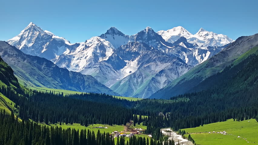 Aerial view of snow-capped peaks and lush green forest in Tianshan Mountains, Xinjiang, China.