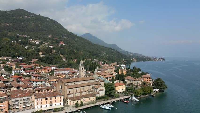 Drone pullback from Duomo di Salo on Lake Garda, drifting backward over the waterfront promenade with moored boats and terracotta roofs as green hills rise behind the town in bright summer sunlight under a calm blue sky
