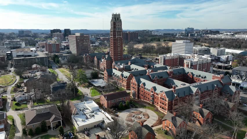 Sunny aerial flyover of Vanderbilt University in Nashville, Tennessee, showing historic red-brick academic buildings and iconic Kirkland Hall tower within private campus setting. Wide shot. Blue sky.