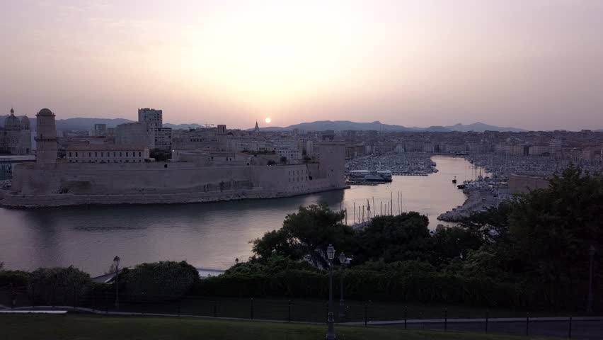Wide view of Marseille Old Port at sunset featuring Fort Saint Jean, calm harbor water, marina filled with boats, and city skyline under warm evening light in southern France