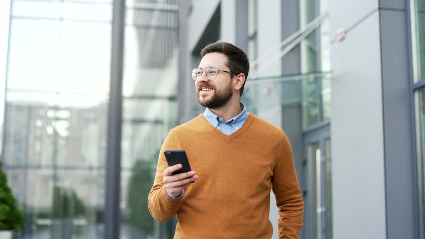Smiling businessman walking on street holding smartphone outside modern office building. Confident handsome bearded entrepreneur in sweater and glasses going to work checking phone in urban city