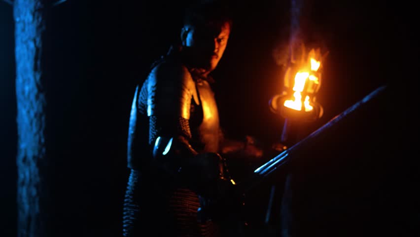 A knight in full armor, his face bloodied and scarred, holding a sword and torch, is sneaking through a dark night forest. A medieval warrior searches in the darkness.