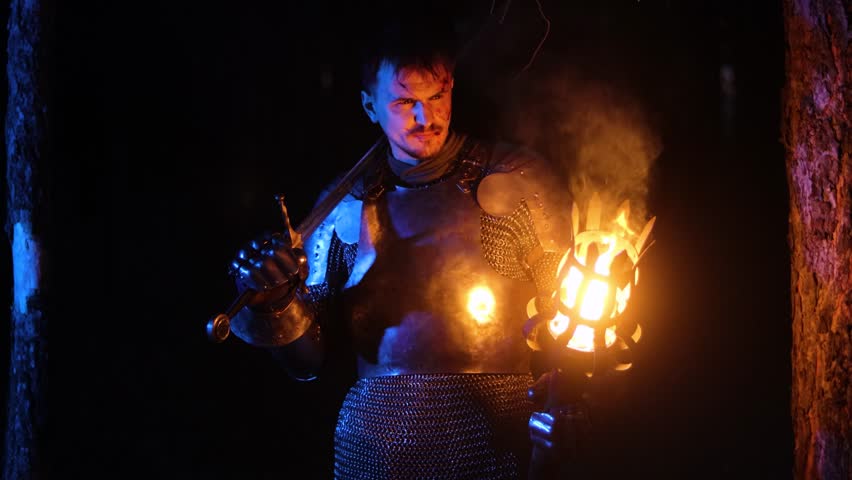 A knight in full armor, his face bloodied and scarred, holding a sword and torch, is sneaking through a dark night forest. A medieval warrior searches in the darkness.