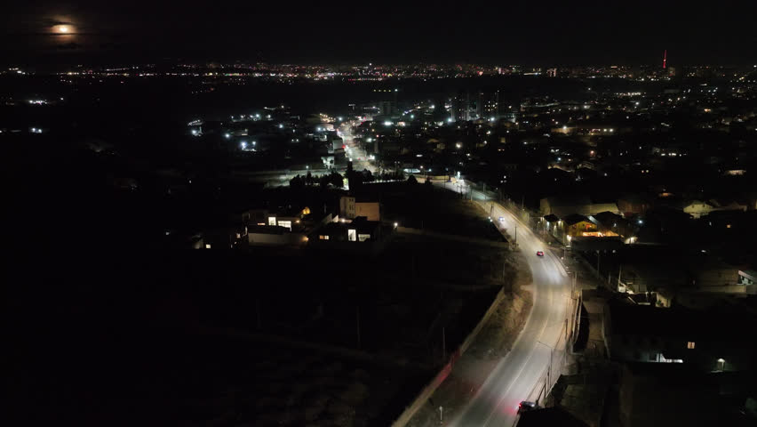 Aerial night view of a winding road with glowing car lights. City lights stretch into the horizon under a dark sky. Traffic moves steadily along the illuminated highway. Scene shows urban expansion