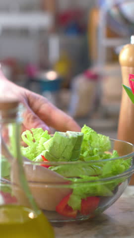 Vertical video. Seasonal salad with oil. A man stirs chopped vegetables in a bowl with a wooden spoon. The process of preparing a healthy salad. Close-up of the food. A cozy kitchen.