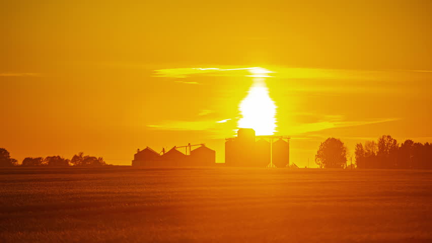 A beautiful orange sunset over farmland and a storafe facility for grain.