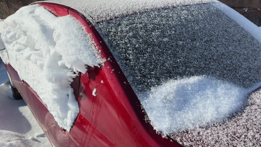 Texture of the snow, settled on the side, rear and front parts of a parked snowbound red car after a snowstorm, view close-up while moving forward in winter sunny day
