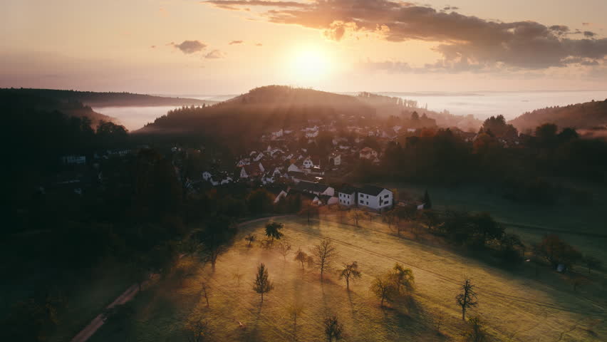 Flying over a rural landscape with hills, meadows and woods, with a village and illuminated fog at sunrise. Aerial footage with sideways motion.
