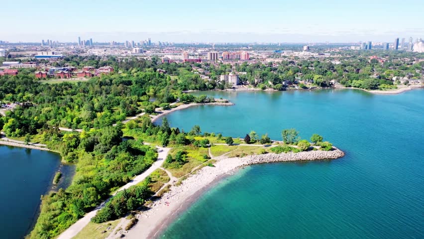 Aerial drone view of Colonel Samuel Smith Park in Etobicoke, Canada, overlooking Lake Ontario with the Toronto city skyline visible in the distance. The scenic waterfront park features green spaces, walking trails, and calm lake waters, creating a peaceful contrast between urban cityscape and natural shoreline landscape.