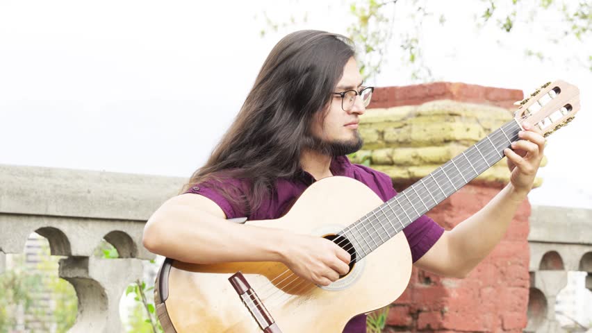 Horizontal medium shot of a male musician with long dark hair and glasses, wearing a purple shirt. He is captured in a moment of deep concentration while playing a classical acoustic guitar.