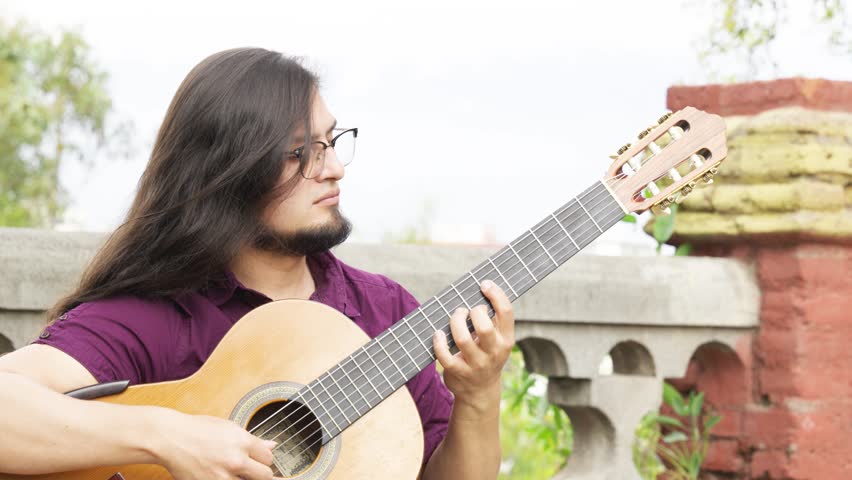 Horizontal close-up shot of a male musician with long dark hair and a goatee, wearing a purple shirt and glasses. He is looking down at the fretboard of his classical acoustic guitar with a look of deep concentration.