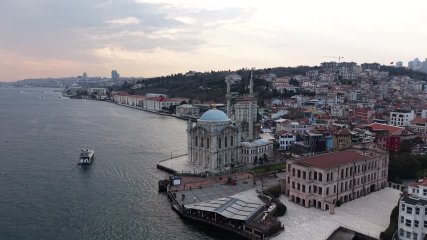 This 4K drone video shows Ortakoy Mosque on the shoreline of Istanbul, Turkiye together with a broader coastal and neighborhood setting. As the camera moves forward, the mosque, seaside square, surrounding historic buildings, pier area, and Bosphorus edge come together within one composition. A ferry moving across the water adds life to the scene, while the hillside settlement and layered urban fabric in the background create a strong city atmosphere. Ortakoy Mosque is the main focal point in this footage, while the waterfront life of Istanbul, Turkiye is also clearly visible. Ortakoy Mosque and the waterfront setting are among the strongest searchable elements here.