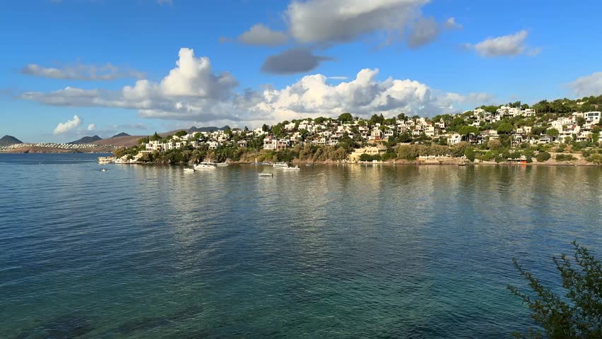 A stunning panoramic view of a tranquil blue bay with elegant white villas nestled on a sunlit hillside under a vast blue sky with soft, drifting white clouds in a Mediterranean setting.