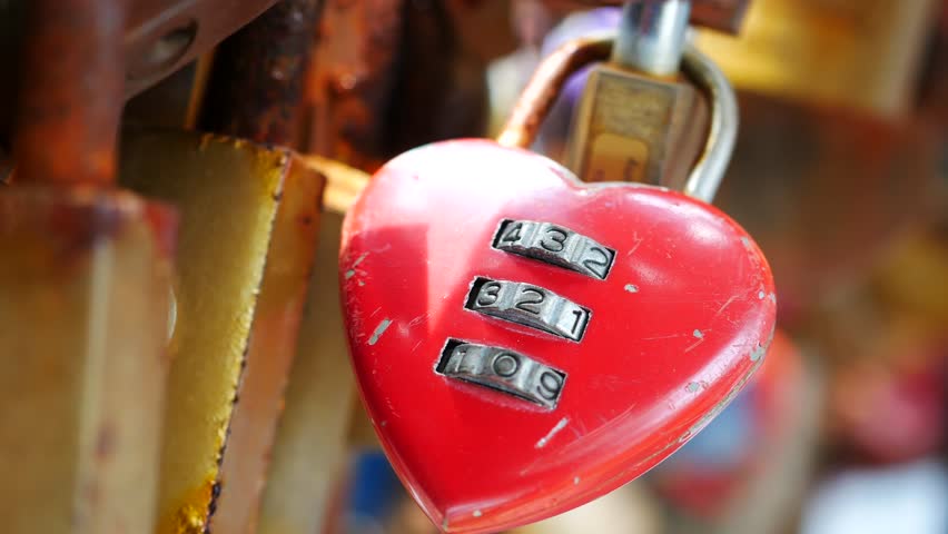 Close up shot of a vibrant red heart-shaped padlock with a three-digit combination lock, symbolizing eternal love and commitment, affixed to a structure with other locks creating a romantic and enduring display