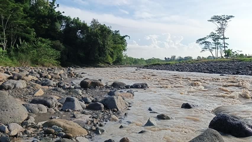 Natural river flowing over rocks with green hills, trees, and blue sky in a peaceful rural landscape environment.