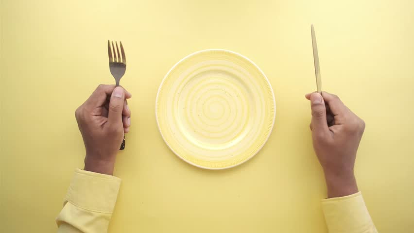 Top view of person holding fork and knife over empty yellow plate on yellow background