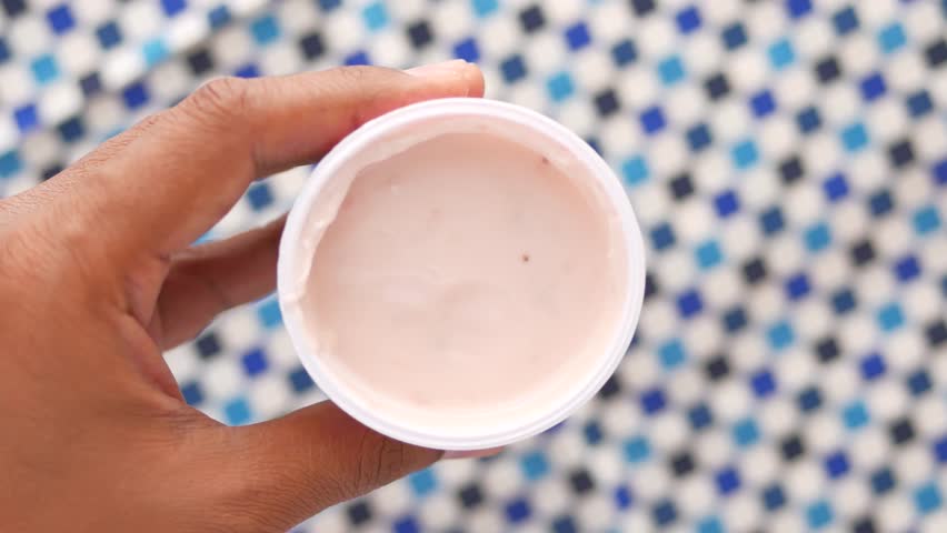 Person scooping pink strawberry yogurt with spoon from white container top view on blue patterned surface