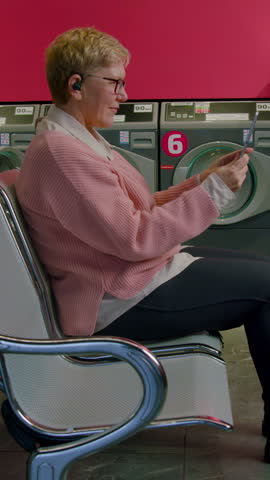 Senior woman in pink cardigan sits in laundromat chair, holding laundry items while waiting for washing machine cycle to complete in a bright, modern setting