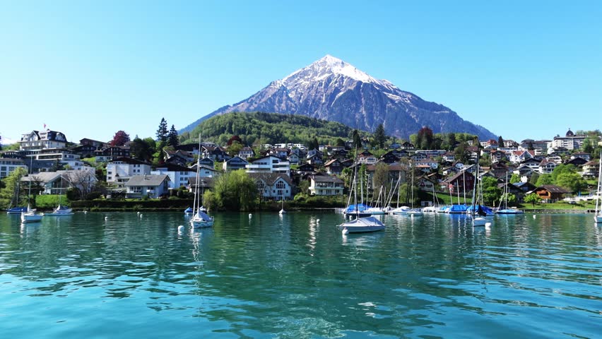 Spiez Schiffstation n snow-capped mountain with white snow peak or swiss alps over house village city on calm water wave with boat ship or cruise ferry on blue sky, 4k Lake Thun Interlaken Switzerland