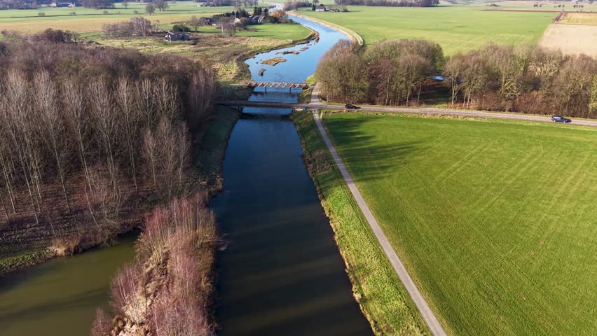 An aerial drone view of a calm canal cutting through a vibrant green rural landscape, featuring a small bridge, a parallel cycling path, and late winter trees under soft sunlight.