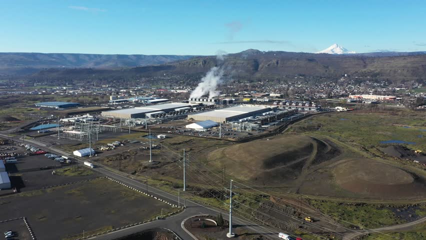 4K aerial drone footage of a large hyperscale data center facility with cooling steam rising from industrial cooling systems in The Dalles, Oregon, USA. Massive server buildings and nearby electrical infrastructure highlight the huge energy demand required for cloud computing, artificial intelligence processing and global internet data storage. Mount Hood is visible in the distance behind the modern technology campus.