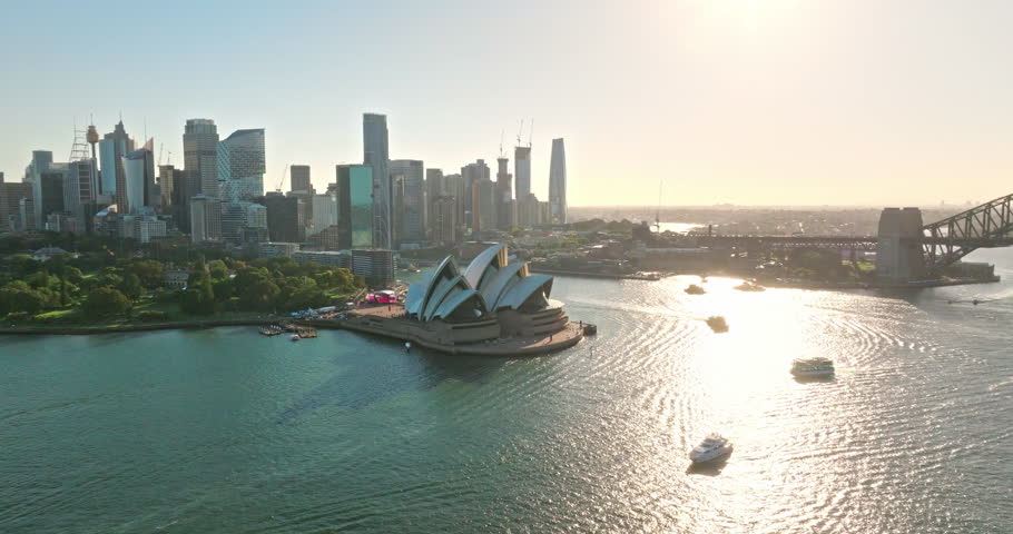 Aerial panorama of Sydney Opera House and Circular Quay waters with ferries and boats, framed by modern CBD skyline under bright blue sky on a warm, sunny day bustling with activity. Explore Australia